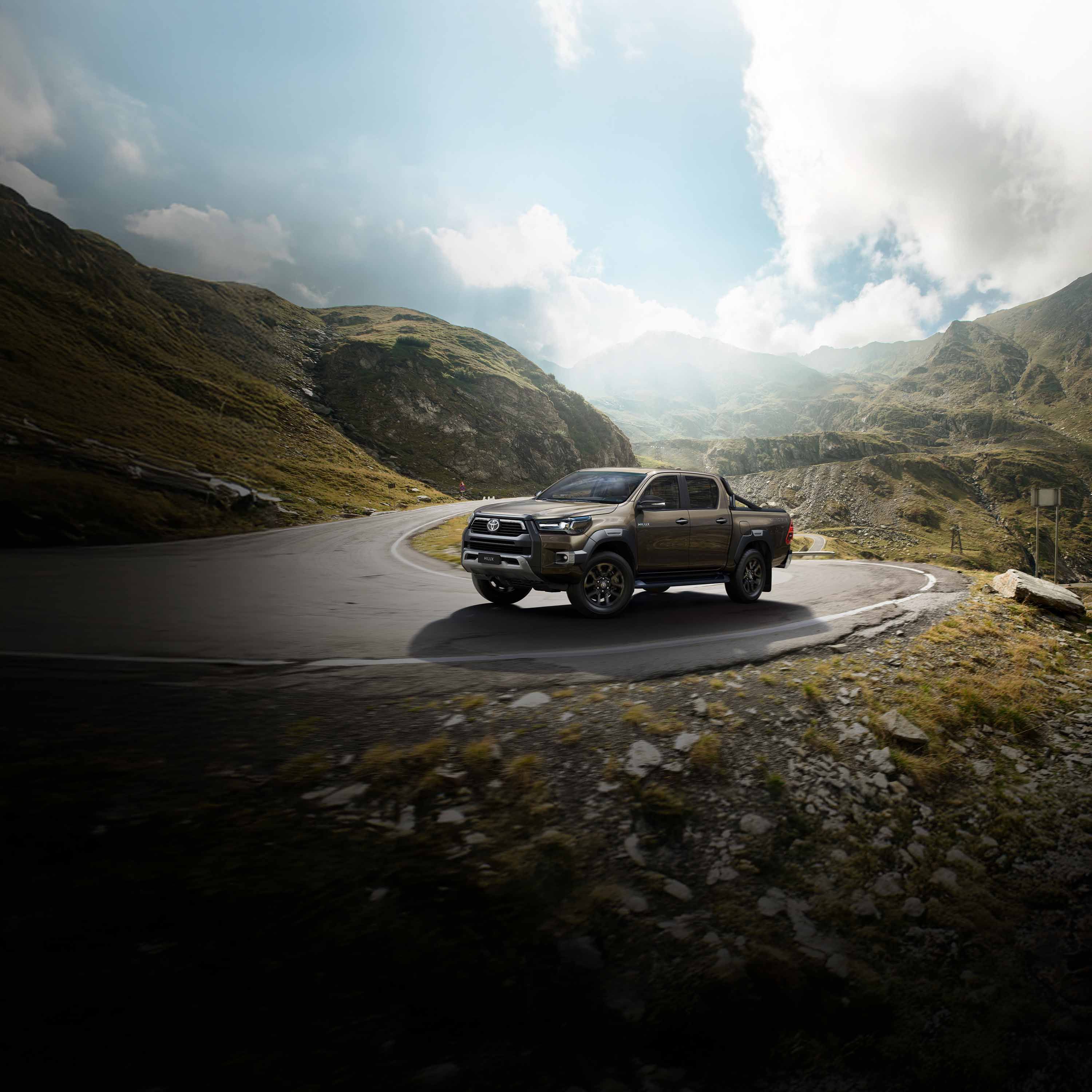 Brauner Pickup fährt Serpentinenstraße entlang in bergiger Landschaft.