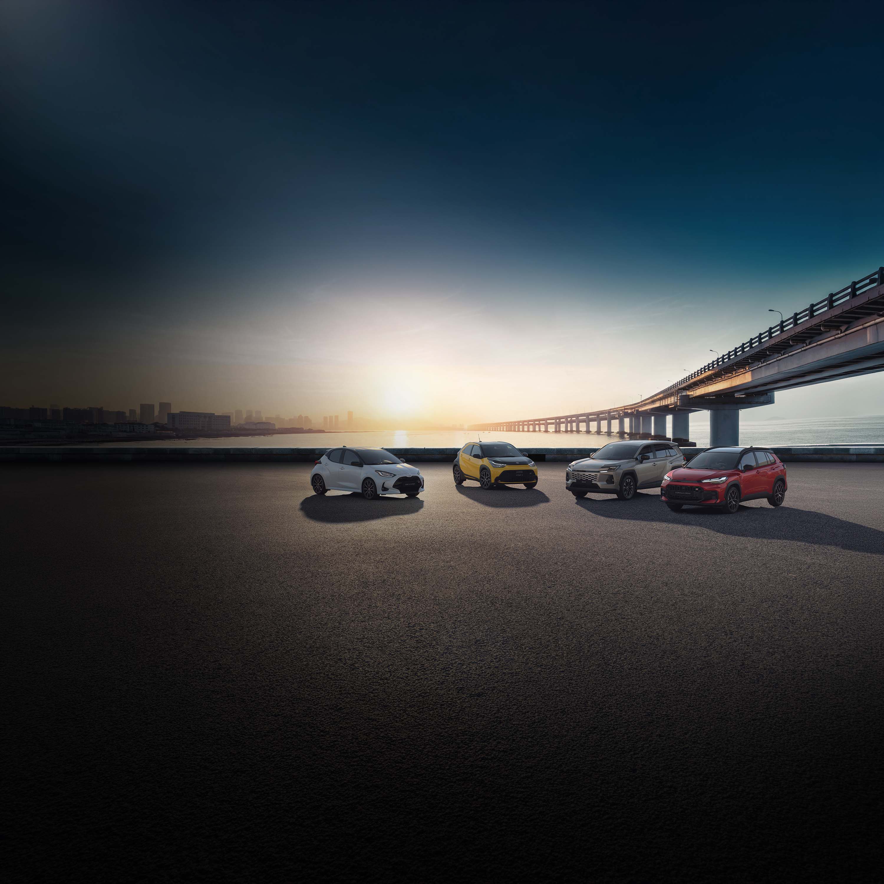 Vier Toyota Autos stehen nebeneinander auf einer offenen Fläche unter einer langen Brücke bei Dämmerung