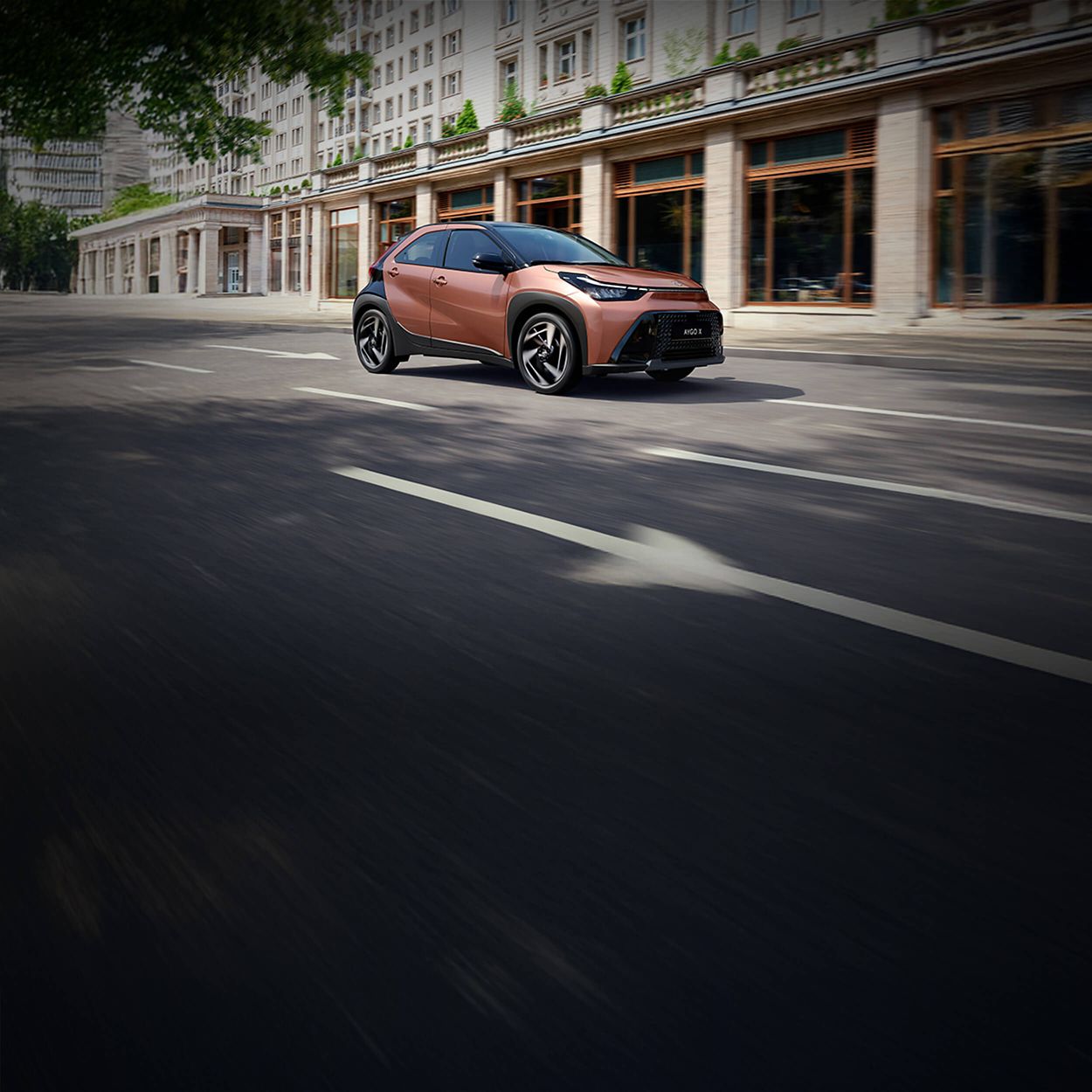 Bronzefarbener Kleinwagen Toyota Aygo X fährt durch eine Innenstadt vor einem Gebäude mit Glasfassade.