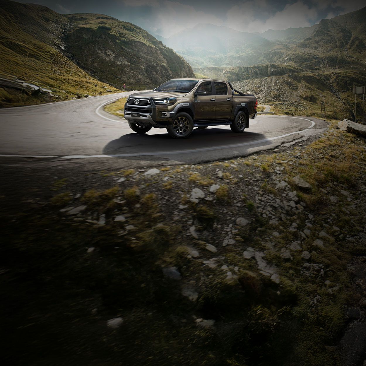 Brauner Pickup fährt Serpentinenstraße entlang in bergiger Landschaft.