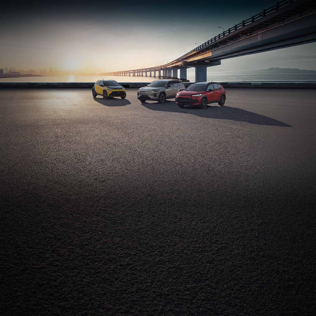 Drei Toyota Autos in Gelb, Grau und Rot stehen auf einem großen, leeren Asphaltplatz unter einer langen Brücke bei Sonnenuntergang