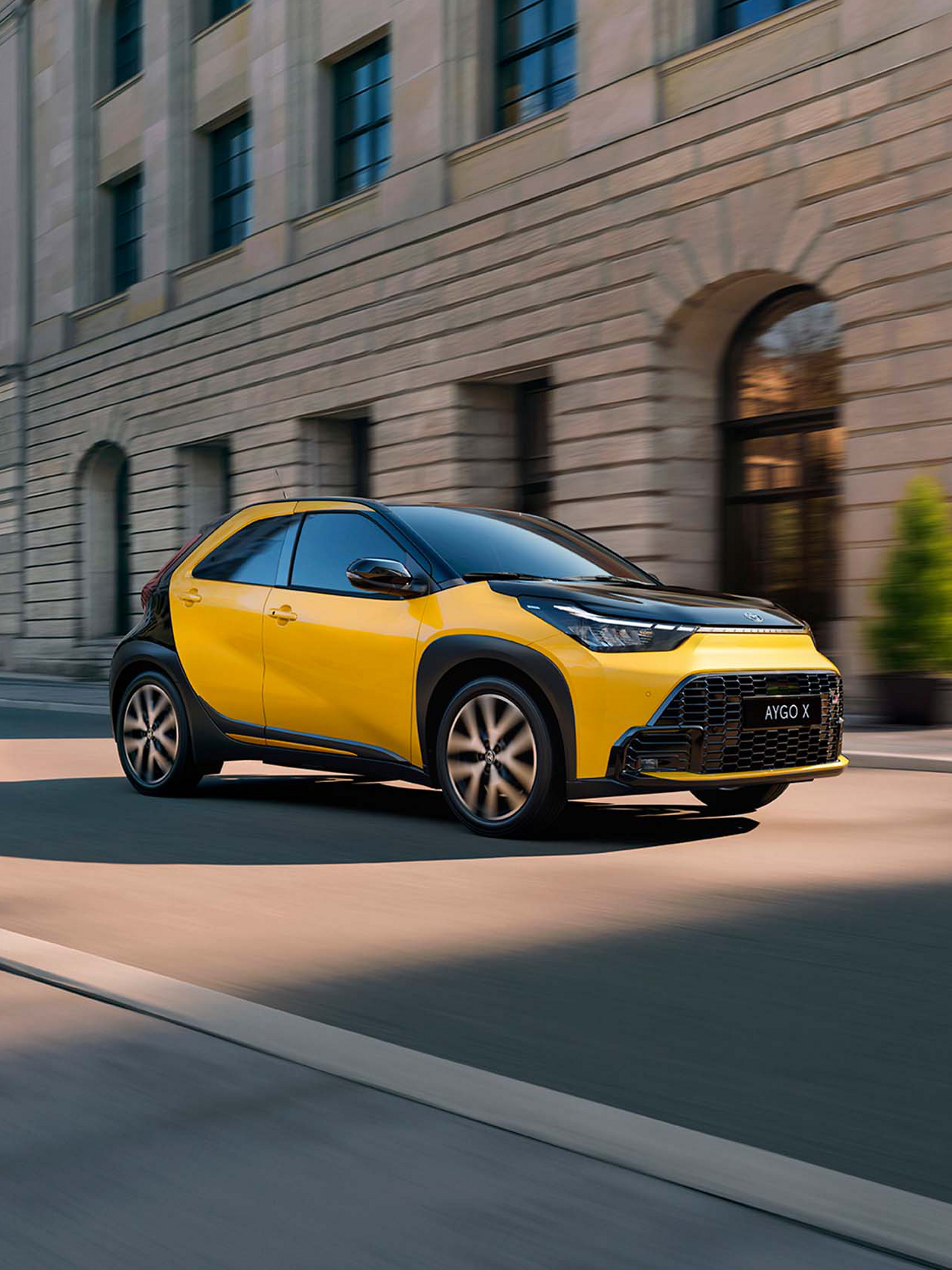 Gelber Toyota Aygo X im Stadtverkehr vor moderner Architektur.