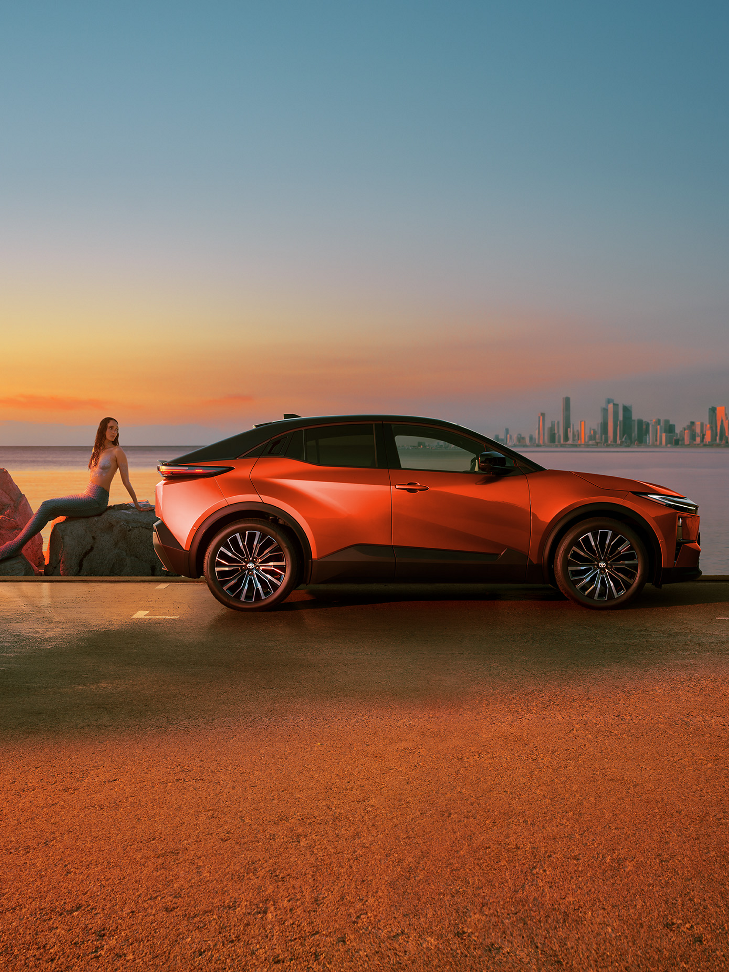 Ein orangefarbener C-HR+ steht bei Sonnenuntergang an einer Küstenstraße; eine Person sitzt entspannt auf Felsen daneben, im Hintergrund sind Meer und eine Skyline zu sehen.