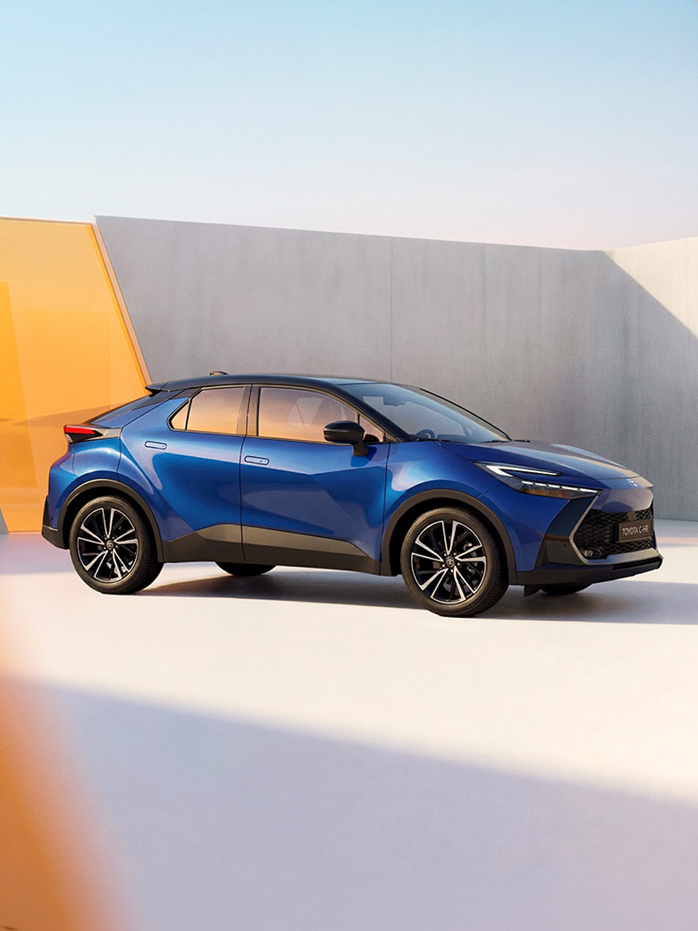 Blauer Toyota C‑HR, seitlich aufgenommen, mit schwarzem Dach und dunklen Felgen, auf einer glatten Fläche vor einer minimalistischen Wand in Grau mit orangefarbener geometrischer Fläche im Hintergrund.