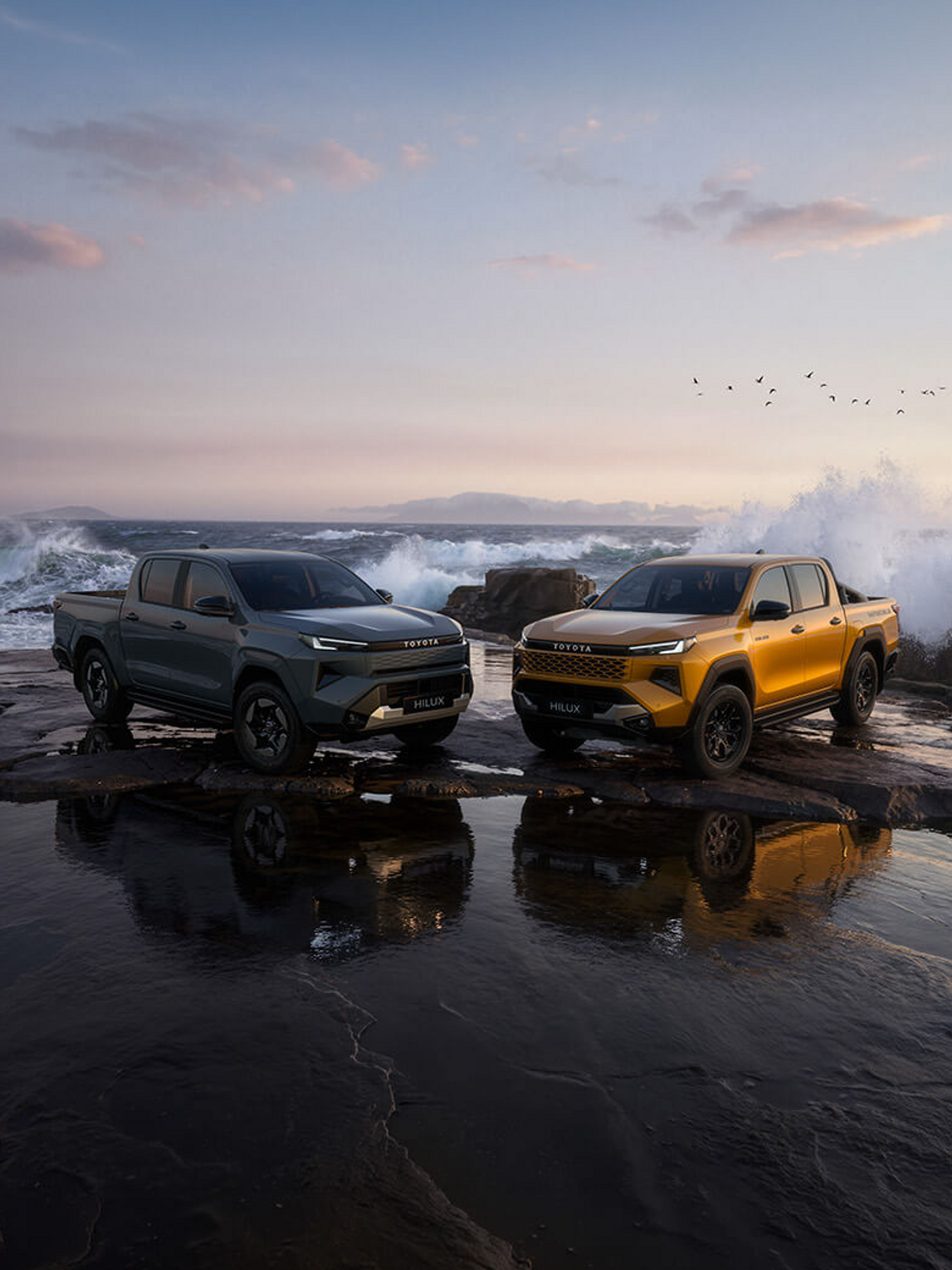 Zwei Toyota Hilux stehen auf einer felsigen Küste vor dem Meer, während Wellen im Hintergrund gegen die Felsen schlagen.