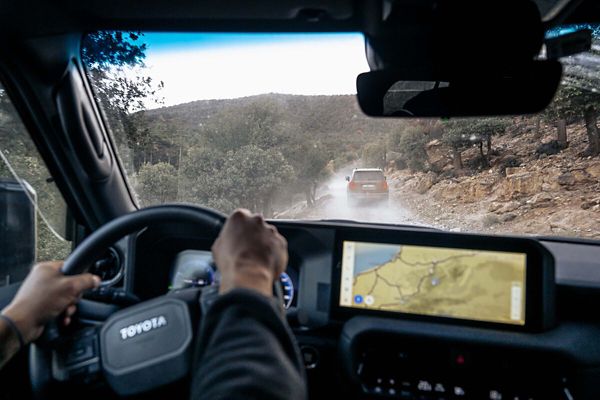 Blick auf das Cockpit des Toyota Land Cruiser und die Hände eines Fahrers am Steuer, voraus fährt auf einer staubigen Piste ein weiterer Land Cruiser