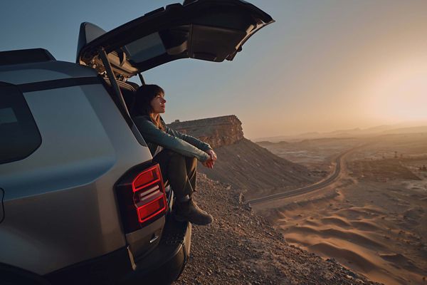 Eine junge Frau sitzt im geöffneten Kofferraum eines am Abhang geparkten grauen Land Cruiser und blickt auf eine weite Wüstenebene im Sonnenuntergang 