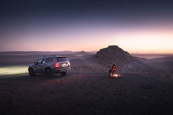 Eine junge Frau in der Dämmerung unter Sternenhimmel an einem Lagerfeuer in der Wüste, daneben ein grauer Land Cruiser mit eingeschalteter Beleuchtung