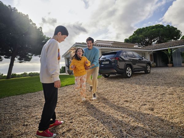 Eine Frau mit zwei spielenden Kindern steht vor der Einfahrt eines Einfamilienhauses mit grünem Garten, in dessen Einfahrt ein Toyota geparkt ist.
