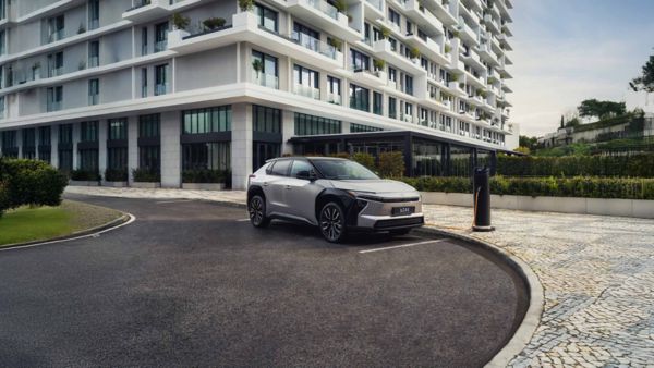 Ein silberner Toyota bZ4X steht auf einem Parkplatz vor einem modernem Bürogebäude und lädt an einer Ladesäule.