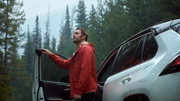 Mann steigt aus der Fahrerseite seines weißen Wagens aus und hat die Fahrertür in der rechten Hand, während er in die Ferne schaut.