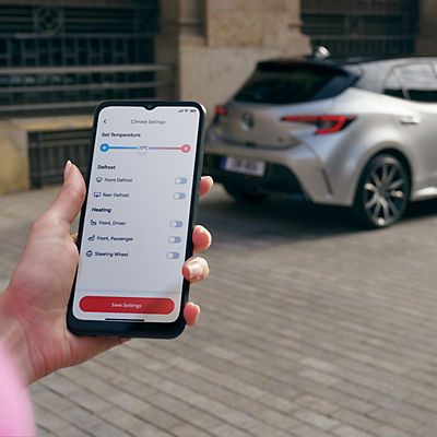 Person mit Smartphone in der Hand steht vor einem geparkten Toyota. Auf dem Bildschirm ist die MyToyota App geöffnet auf der die Temperatur angezeigt wird.