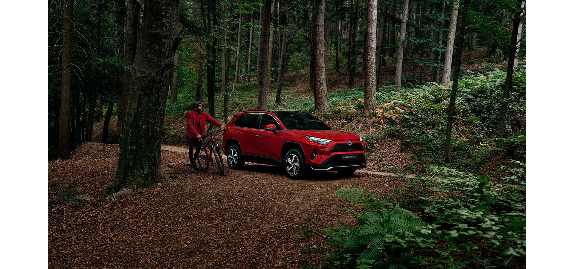 Man in red jacket, stood with a bike next to a red RAV4, in a forest