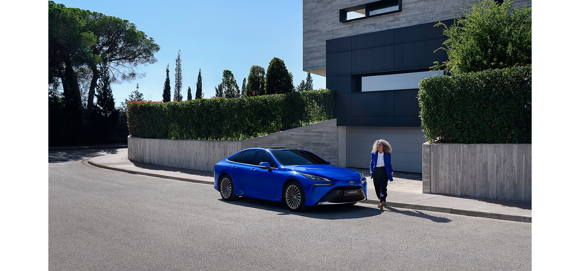 Woman stepping out of a modern house and walking around a bright blue Toyota Mirai.