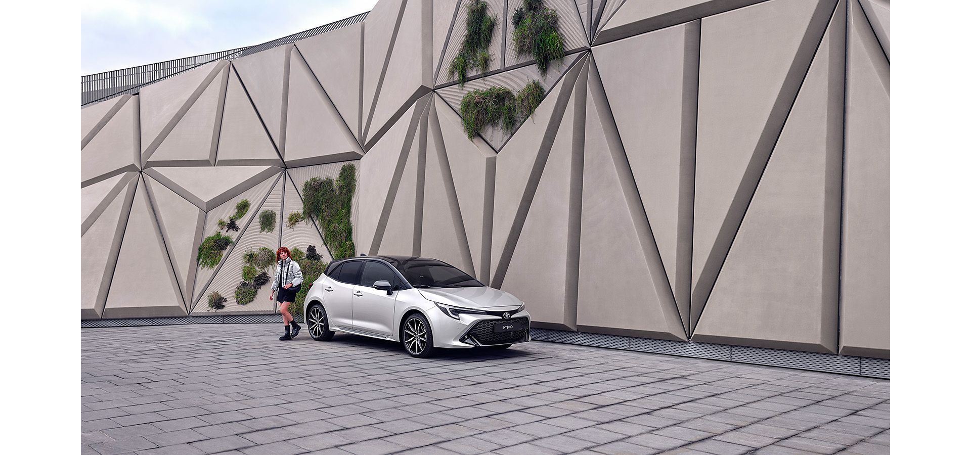 Woman walking outside of a modern abstract building, walking around a silver Corolla Hatchback
