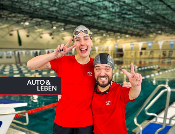 Zwei lächelnde Schwimmer in roten T-Shirts posieren zusammen in einem Schwimmbad, wobei einer ein Peace-Zeichen macht und der andere den Daumen nach oben zeigt