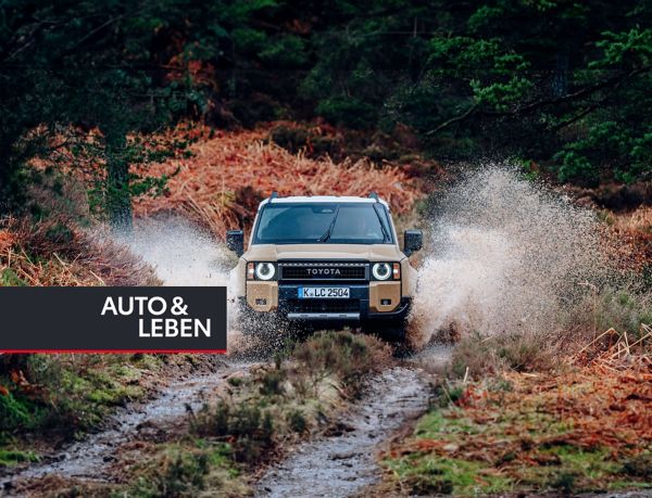 Ein beiger Geländewagen fährt durch einen schlammigen Waldweg, spritzt Wasser um sich herum, umgeben von üppigen grünen Bäumen und Herbstlaub.