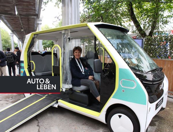 Eine Frau sitzt an einem belebten Platz in einem kleinen, seitlich offenen Shuttle-Bus mit ausgefahrener Rollstuhlrampe zum hinteren Fahrgastbereich