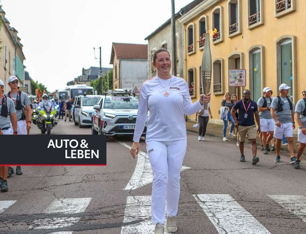 Carina Hoffmann, weiß gekleidet, mit einer goldenen Fackel in der Hand, eskortiert von offiziellen Begleitern, im Hintergrund Fahrzeuge und Zuschauer