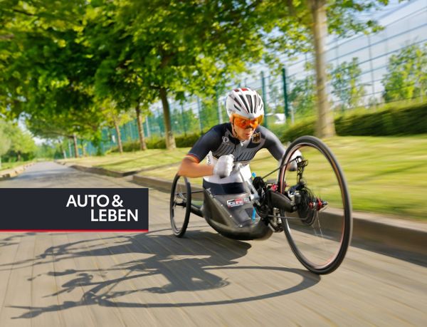 Paralympics-Sportlerin während der Fahrt in ihrem Handbike schräg von vorne dynamisch fotografiert, im Hintergrund Bäume und ein Sportgelände