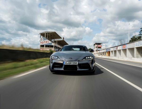 Ein grauer GR Supra von vorn fotografiert auf einer leeren Rennstrecke fahrend, im Hintergrund eine alte Tribüne und die Boxengasse mit Werbetafeln