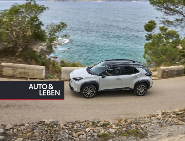 Ein hellgrauer Yaris Cross mit schwarzem Dach auf einer Küstenstraße, im Vordergrund Geröll, im Hintergrund das Meer und mediterrane Bäume