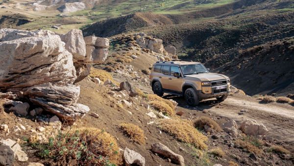 Ein sandfarbener Toyota Land Cruiser fährt auf einem unbefestigten Weg durch eine karge Gebirgslandschaft, im Vordergrund Felsen auf einer Anhöhe