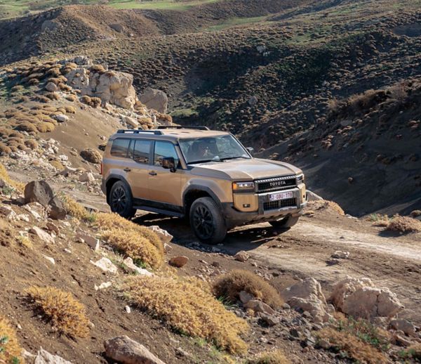 Ein sandfarbener Toyota Land Cruiser fährt auf einem unbefestigten Weg durch eine karge Gebirgslandschaft, im Vordergrund Felsen auf einer Anhöhe