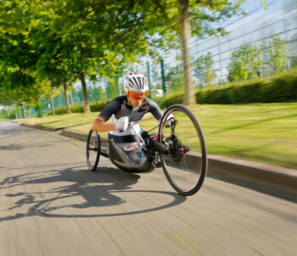 Paralympics-Sportlerin während der Fahrt in ihrem Handbike schräg von vorne dynamisch fotografiert, im Hintergrund Bäume und ein Sportgelände