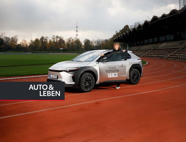 Ulrike Nasse-Meyfarth steigt in einen silbergrauen Toyota bZ4X, der auf der Laufbahn eines kleinen Stadions steht, im Hintergrund die Tribüne und Rasen