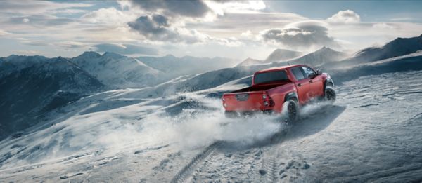 Ein roter Toyota Hilux fährt einen steilen, verschneiten Berg hinauf, während im Hintergrund hohe Berge von der Sonne angestrahlt werden