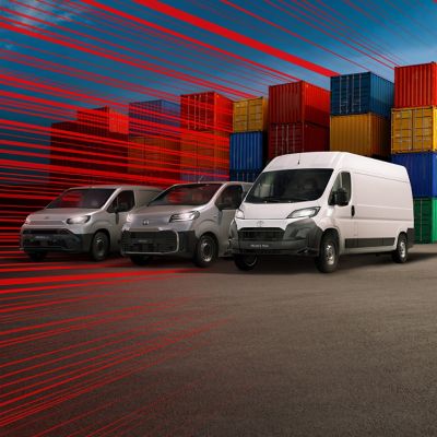 Proace Max, Proace und ein Proace City schräg von vorn auf einem Parkplatz, umgeben von Stapeln aus Europaletten, daneben Ladesäulen