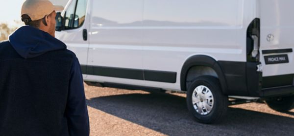 Eine Person mit einer Mütze steht vor einem weißen Toyota Proace Max, der auf einer Schotterstraße geparkt ist.