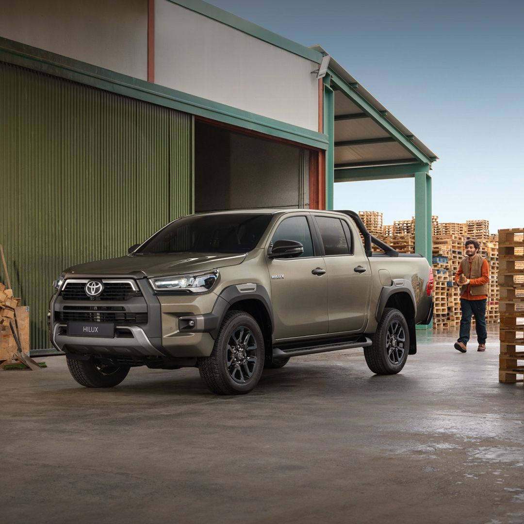 Ein grau-brauner Toyota Hilux steht vor einer Lagerhalle, daneben stehen Holz-Paletten.