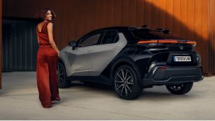 Eine Frau in kastanienbraunem Oberteil geht auf einen geparkten,schwarzen Toyota C-HR zu