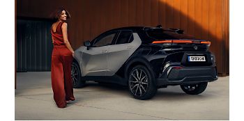 Eine Frau in kastanienbraunem Oberteil geht auf einen geparkten,schwarzen Toyota C-HR zu
