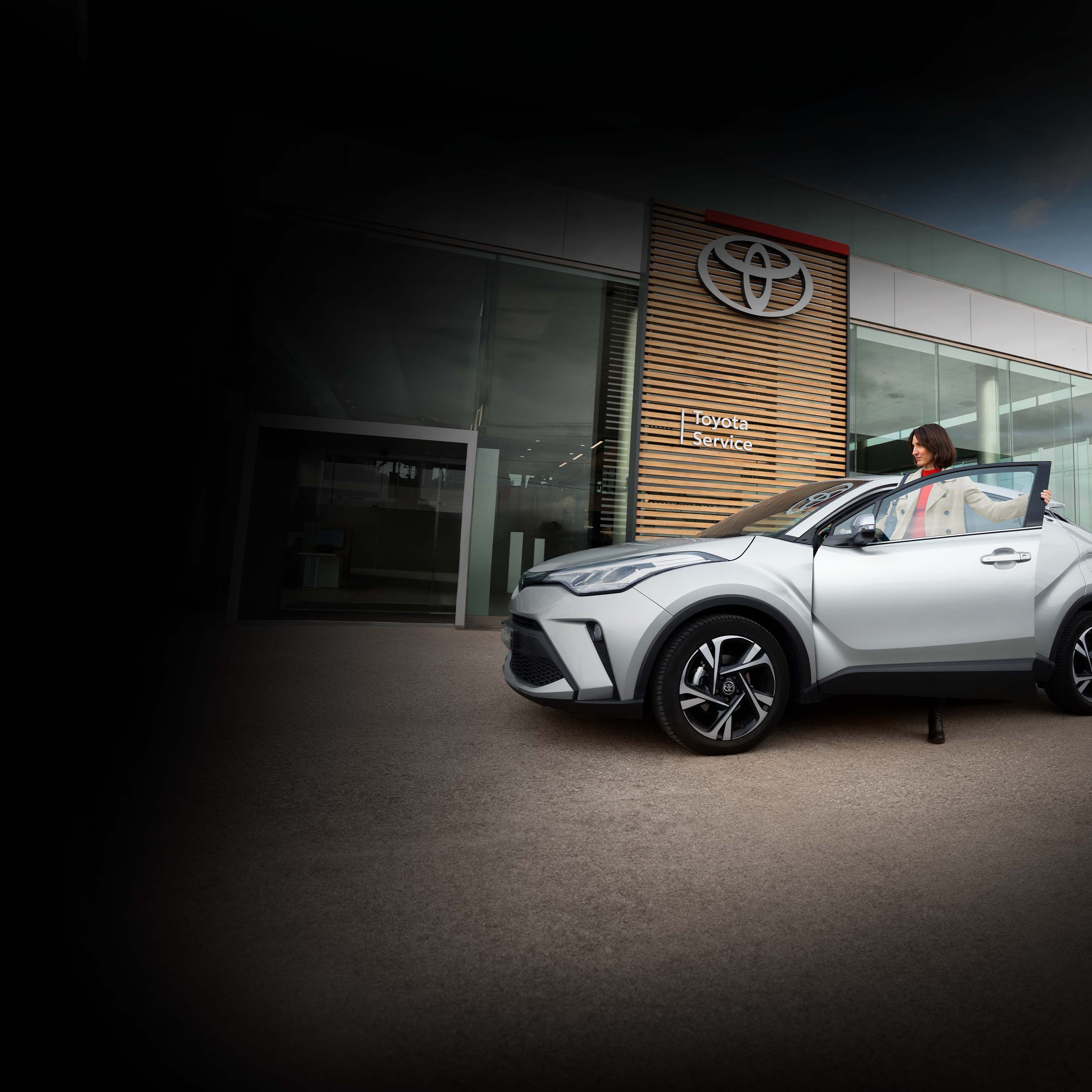 Eine Frau steigt in den Fahrersitz eines silbernen Autos. Im Hintergrund ist ein Autohauseingang, an dessen Wand die Aufschrift Toyota Service steht.