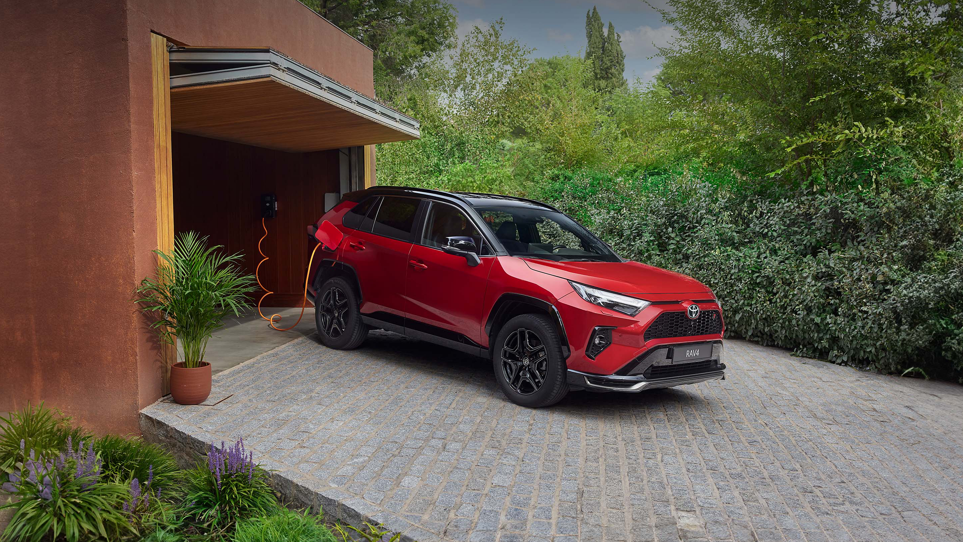 Roter Toyota RAV4 als Plug-in Hybrid steht vor einer Garageneinfahrt und lädt an einer Wallbox.