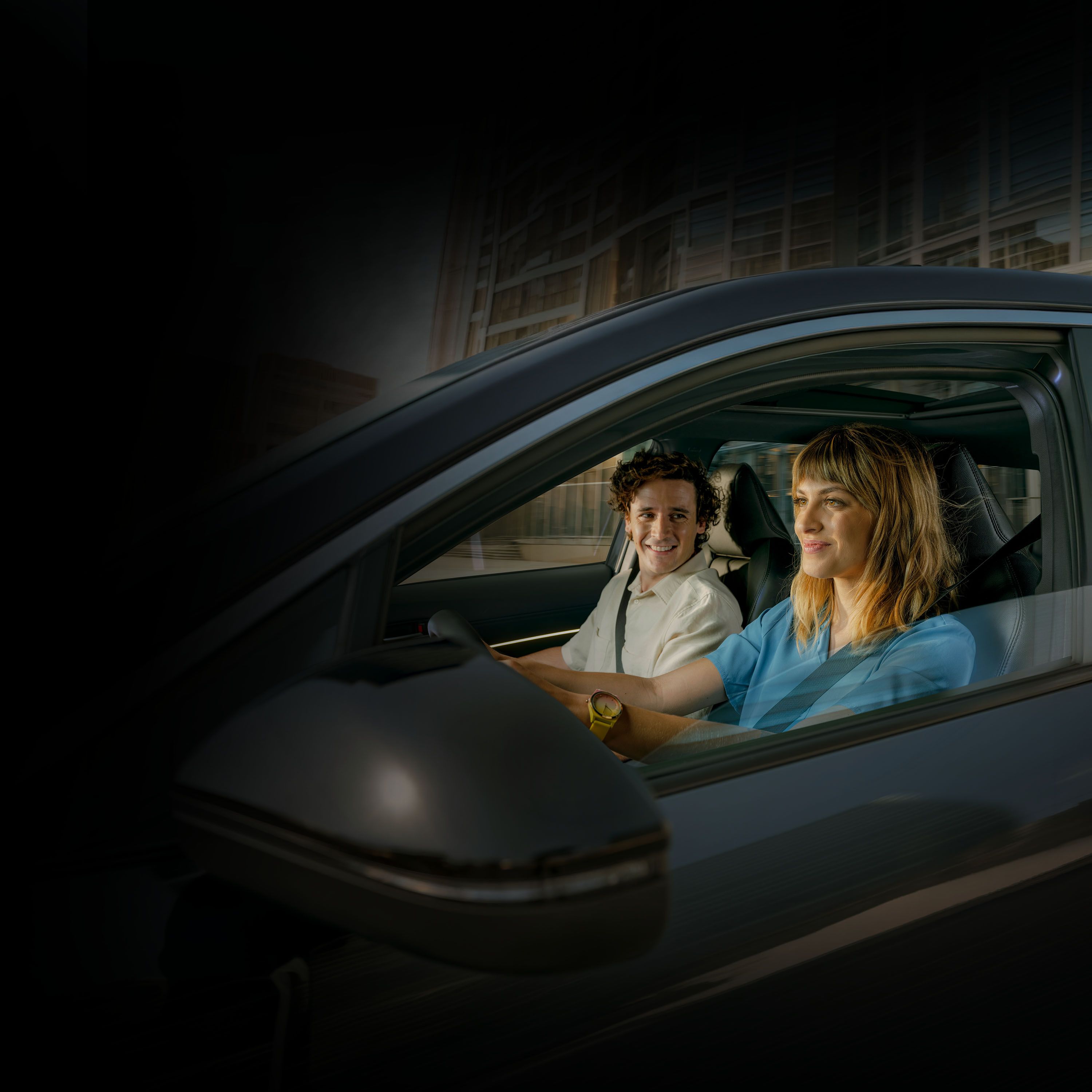 Seitenansicht eines Toyota mit einer Fahrerin und einem Beifahrer im Innenraum. Beide tragen Sicherheitsgurte und fahren durch eine nächtliche Stadt.