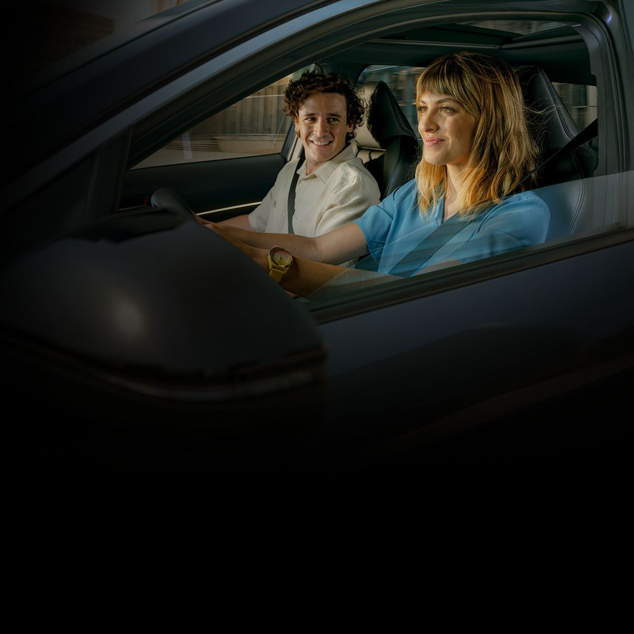 Seitenansicht eines Toyota mit einer Fahrerin und einem Beifahrer im Innenraum. Beide tragen Sicherheitsgurte und fahren durch eine nächtliche Stadt.