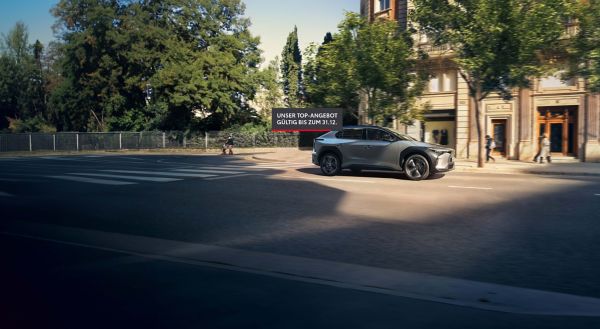 Grauer Toyota bZ4X fährt auf einer Straße in der Innenstadt im Sonnenschein, darauf ein Banner mit der Aufschrift "Unser Top Angebot zum 31.12."