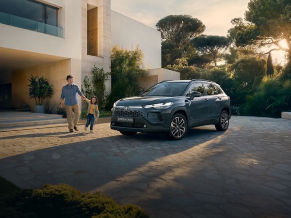 Ein Corolla Cross steht in der Einfahrt eines großen modernen Familienhauses, während ein Mann mit seinem Kind in der Hand darauf zuläuft.