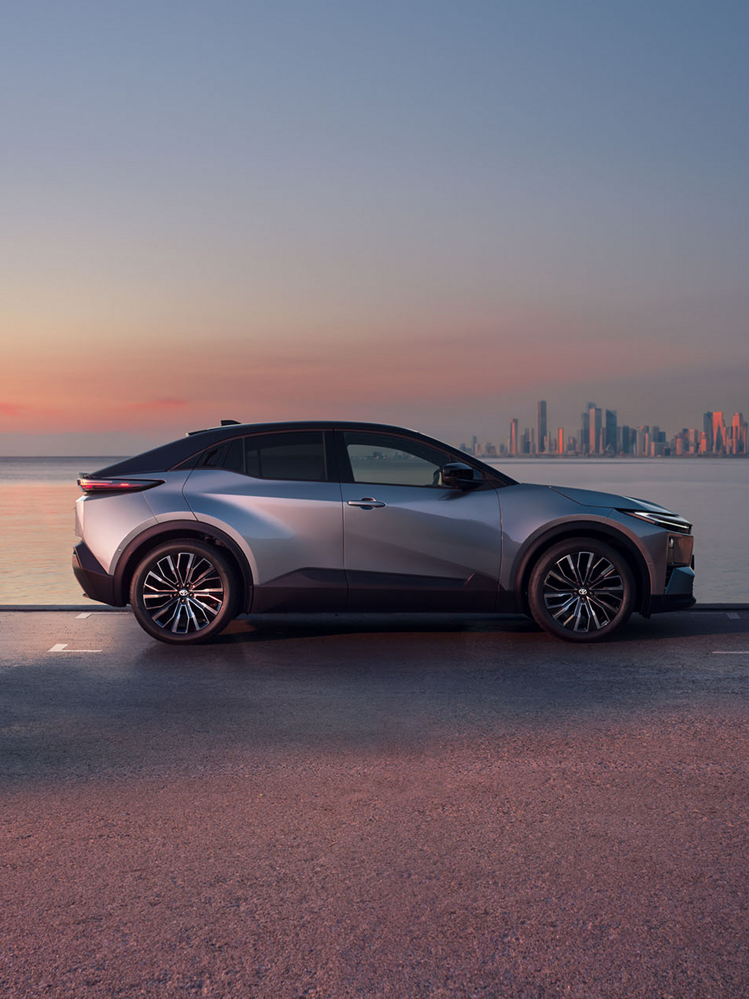 Ein silberner Toyota C-HR+ seitlich vor der Skyline einer großen Stadt mit Wolkenkratzern im Sonnenuntergang