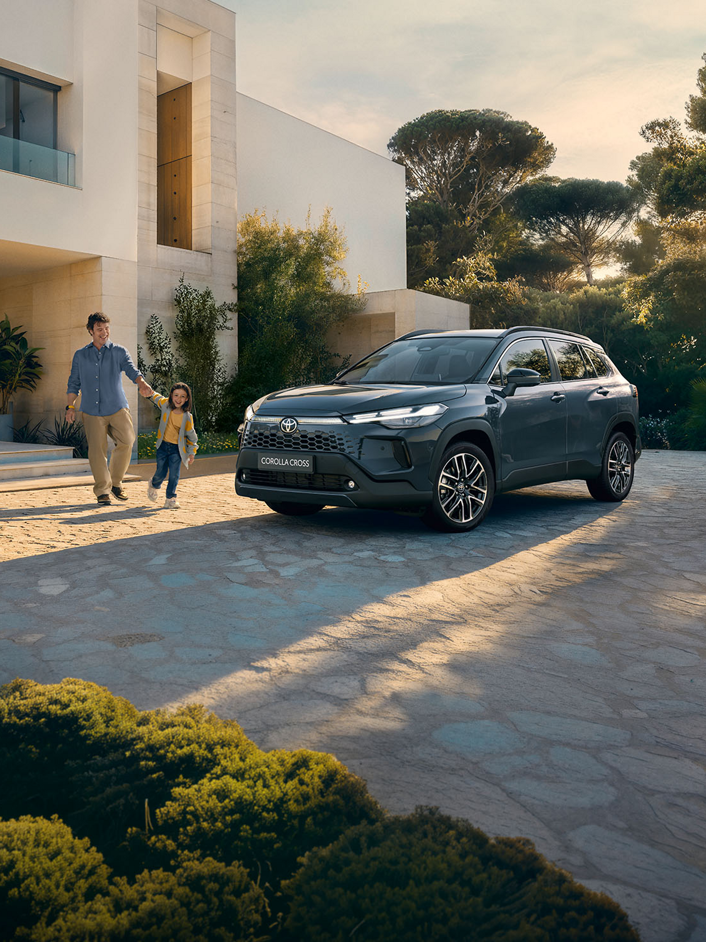 Ein Corolla Cross steht in der Einfahrt eines großen modernen Familienhauses, während ein Mann mit seinem Kind in der Hand darauf zuläuft.