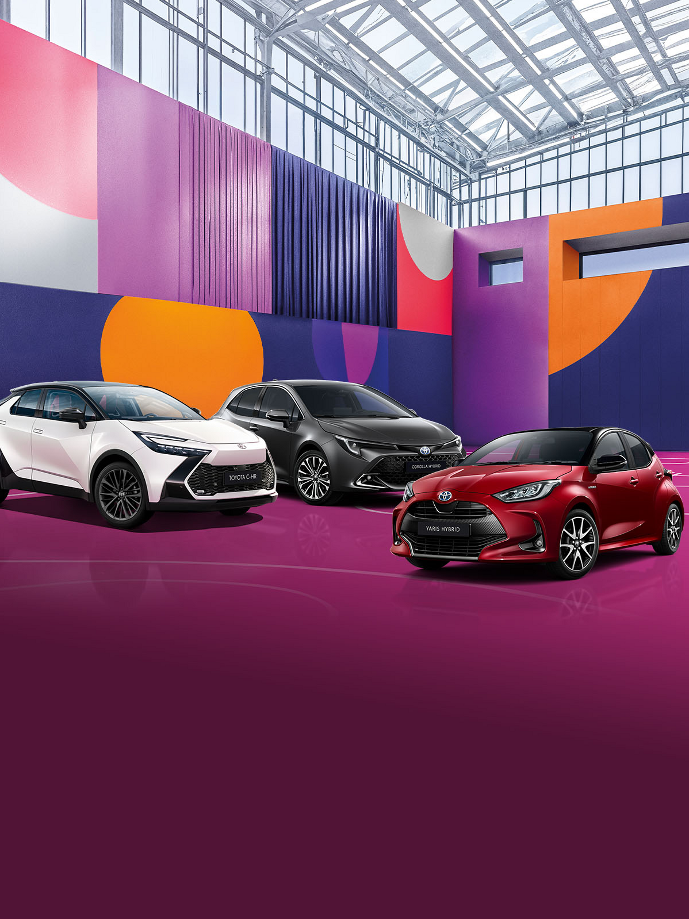 Ein weißer Toyota C-HR steht neben einem schwarzen Corolla und roten Yaris auf einem Basketballplatz, der mit verschiedenen lila Schattierungen und bunten Forman angemalt ist. Darüber ist ein Glasdach.