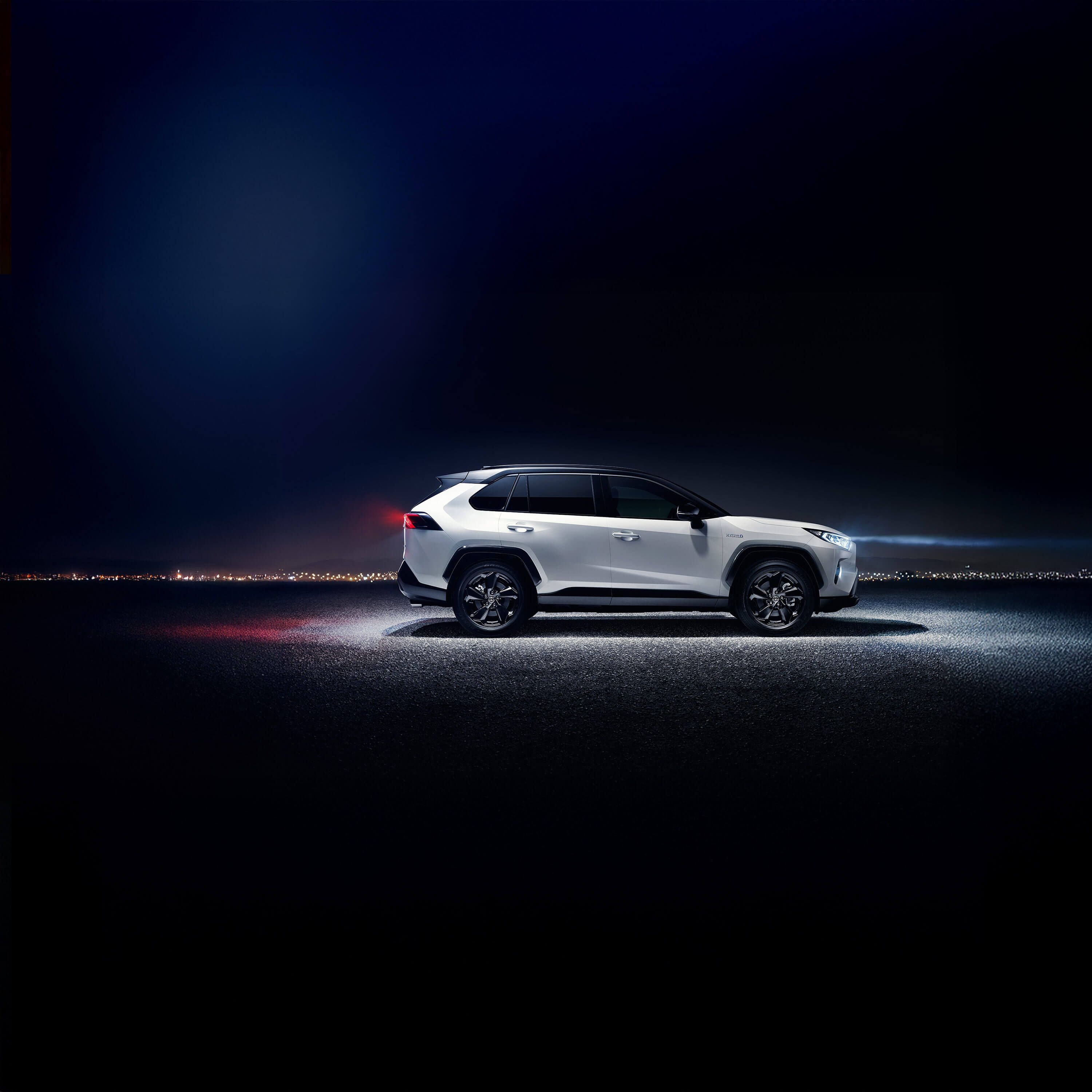 Seitenansicht eines weißen Toyota RAV4 SUV, der bei Nacht im Scheinwerferlicht auf einer asphaltierten Straße steht mit Gebäudelichtern in der Ferne.