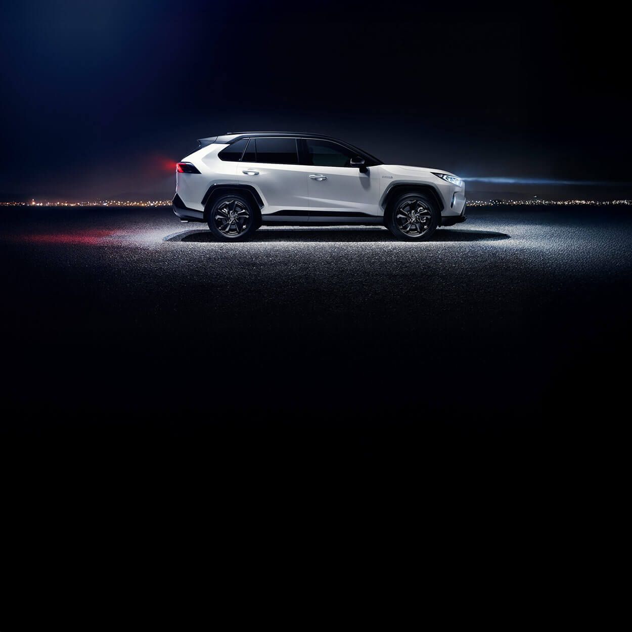 Seitenansicht eines weißen Toyota RAV4 SUV, der bei Nacht im Scheinwerferlicht auf einer asphaltierten Straße steht mit Gebäudelichtern in der Ferne.