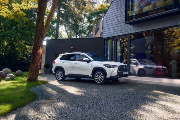 Weißer Mittelklasse-SUV steht geparkt in einer Einfahrt vor einem modernen Haus und vor Sonne geschützt unter Kiefern.