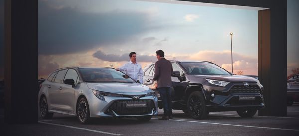 Zwei Personen sind in einem Beratungsgespräch über gebrauchte SUV-Fahrzeuge. Einer davon trägt ein Hemd mit Toyota Logo. Beide stehen zwischen zwei gebrauchten Fahrzeugen.