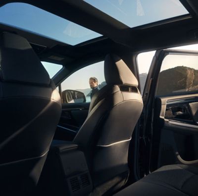 Innenraum eines SUV-Modells mit Panoramadach und schwarzen Ledersitzen. Die Rückbanktür steht offen und am Beifahrerfenster steht ein Mann.